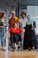 A diverse group of young business people walking a corridor in the glass-enclosed office of a modern startup, including a person in a wheelchair and a woman wearing a hijab, showing a dynamic mix of photo