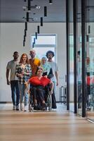 A diverse group of young business people walking a corridor in the glass-enclosed office of a modern startup, including a person in a wheelchair and a woman wearing a hijab, showing a dynamic mix of photo