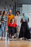 A diverse group of young business people walking a corridor in the glass-enclosed office of a modern startup, including a person in a wheelchair and a woman wearing a hijab, showing a dynamic mix of photo