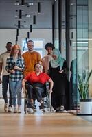 A diverse group of young business people walking a corridor in the glass-enclosed office of a modern startup, including a person in a wheelchair and a woman wearing a hijab, showing a dynamic mix of photo
