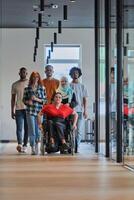 A diverse group of young business people walking a corridor in the glass-enclosed office of a modern startup, including a person in a wheelchair and a woman wearing a hijab, showing a dynamic mix of photo