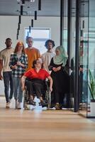 A diverse group of young business people walking a corridor in the glass-enclosed office of a modern startup, including a person in a wheelchair and a woman wearing a hijab, showing a dynamic mix of photo