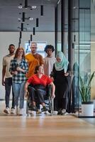 A diverse group of young business people walking a corridor in the glass-enclosed office of a modern startup, including a person in a wheelchair and a woman wearing a hijab, showing a dynamic mix of photo