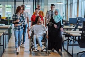A diverse group of young business people walking a corridor in the glass-enclosed office of a modern startup, including a person in a wheelchair and a woman wearing a hijab, showing a dynamic mix of photo