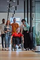 A diverse group of young business people walking a corridor in the glass-enclosed office of a modern startup, including a person in a wheelchair and a woman wearing a hijab, showing a dynamic mix of photo