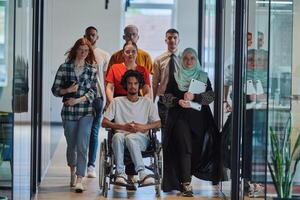 A diverse group of young business people walking a corridor in the glass-enclosed office of a modern startup, including a person in a wheelchair and a woman wearing a hijab, showing a dynamic mix of photo