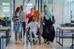 A diverse group of young business people walking a corridor in the glass-enclosed office of a modern startup, including a person in a wheelchair and a woman wearing a hijab, showing a dynamic mix of photo