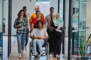 A diverse group of young business people walking a corridor in the glass-enclosed office of a modern startup, including a person in a wheelchair and a woman wearing a hijab, showing a dynamic mix of photo