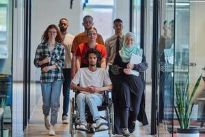 A diverse group of young business people walking a corridor in the glass-enclosed office of a modern startup, including a person in a wheelchair and a woman wearing a hijab, showing a dynamic mix of photo