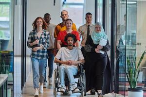 A diverse group of young business people walking a corridor in the glass-enclosed office of a modern startup, including a person in a wheelchair and a woman wearing a hijab, showing a dynamic mix of photo