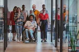 A diverse group of young business people walking a corridor in the glass-enclosed office of a modern startup, including a person in a wheelchair and a woman wearing a hijab, showing a dynamic mix of photo