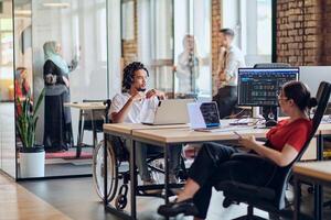 A young business group, including an African American businessman in a wheelchair, collaborates within a modern glass office, actively engaged around a computer and laptop, collectively solving photo