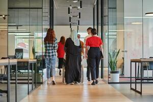 A diverse group of business people walking a corridor in the glass-enclosed office of a modern startup, including a person in a wheelchair and a woman wearing a hijab, showing a dynamic mix of photo
