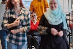A diverse group of business people walking a corridor in the glass-enclosed office of a modern startup, including a person in a wheelchair and a woman wearing a hijab, showing a dynamic mix of photo
