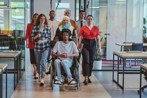 A diverse group of young business people walking a corridor in the glass-enclosed office of a modern startup, including a person in a wheelchair and a woman wearing a hijab, showing a dynamic mix of photo