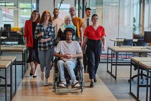 A diverse group of young business people walking a corridor in the glass-enclosed office of a modern startup, including a person in a wheelchair and a woman wearing a hijab, showing a dynamic mix of photo