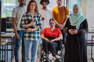 A diverse group of young business people walking a corridor in the glass-enclosed office of a modern startup, including a person in a wheelchair and a woman wearing a hijab, showing a dynamic mix of photo