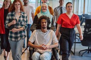 A diverse group of young business people walking a corridor in the glass-enclosed office of a modern startup, including a person in a wheelchair and a woman wearing a hijab, showing a dynamic mix of photo