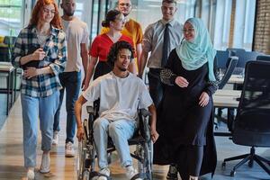 A diverse group of young business people walking a corridor in the glass-enclosed office of a modern startup, including a person in a wheelchair and a woman wearing a hijab, showing a dynamic mix of photo