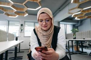 A businesswoman in a hijab using a smartphone in a modern office, epitomizing a successful and empowered professional in today's tech-savvy world photo