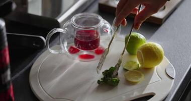 Man Putting Mint Leaves Into Berry Tea With Citrus, Closeup, Glass Teapot, Lemon And Lime At Table video