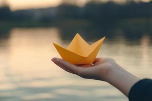 A hand holds a bright yellow paper boat above the surface of tranquil water. photo