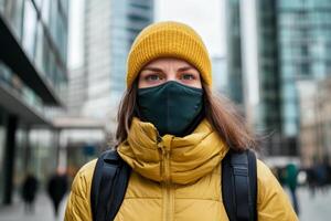Young woman in a mustard yellow jacket and hat stands in an urban setting with a mask amid modern buildings photo