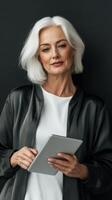 A woman with silver hair and a stylish black jacket stands confidently in front of a dark wall, holding a tablet. She radiates poise and professionalism in a contemporary environment photo