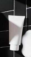 White tube of skincare cream resting against a black tiled wall with foam nearby photo