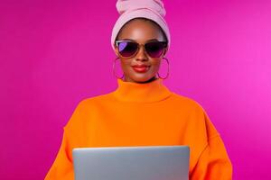 A fashion-forward woman wearing a vibrant orange sweater and pink headscarf types on a laptop, displaying confidence and style. photo