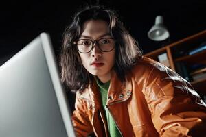 Young person with long hair wearing glasses and jacket engaged in focused computer activity in dimly lit room photo
