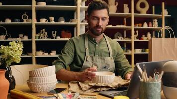 Successful happy Caucasian man male businessman guy small business owner entrepreneur packing wrapping ceramic bowl pottery use calculator calculating price money looking camera smile inside workshop video