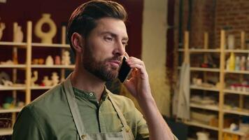 Professional skilled potter Caucasian man male guy businessman small business owner seller potter ceramist artist craftsman talking mobile phone deal call client discussing order in pottery workshop video