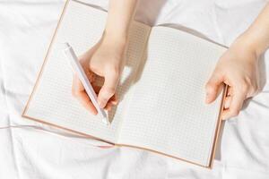 Woman takes notes or makes plan, day results on blank white page of paper in notebook or diary, holds notepad and pen in hands, top view, time for yourself morning or evening before sleep photo