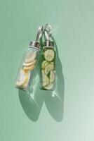 Glass bottles with lemon, cucumber water drink detox at sunlight on green colored background, copy space. Healthy infused water boost metabolism, weight loss. Aesthetic still life, refreshing drink photo