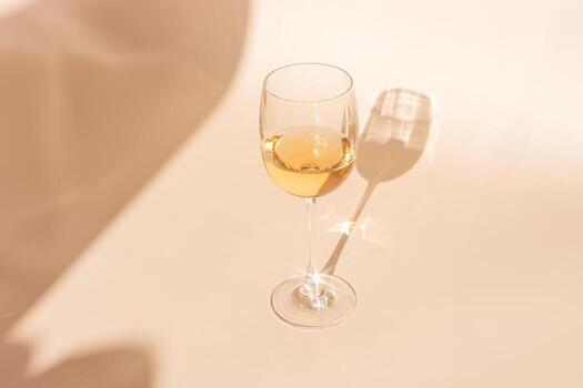 One wineglass with white wine sparkling at sunlight, peach beige background, copy space, dark shadow and reflection, sun flare light effect on table. Alcohol drink top view, neutral pastel photo