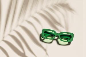 Trendy sunglasses with green Frame on neutral beige background, copyspace, beach vibes, summertime mood, palm leaf shadow, summer fashion vivid glasses. Minimal top view of eyeglasses photo