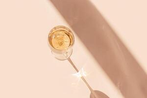 White wine in crystal transparent glass with shadow and glare at sunlight, light white beige background, creative summer alcohol drink minimal concept, copy space, top view, pastel peach color photo