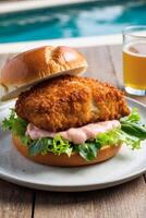 A golden-brown, crispy fish sandwich sits on a bun with lettuce and sauce, next to a refreshing glass of beer on a sunny day by the pool. photo