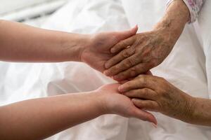 A young womans hands gently hold the hands of an elderly female patient in a hospital bed, symbolizing care, support, and compassion. photo