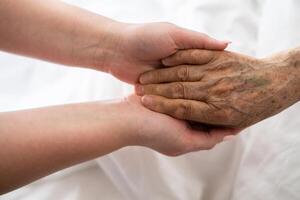 A young womans hands gently hold the hands of an elderly female patient in a hospital bed, symbolizing care, support, and compassion. photo