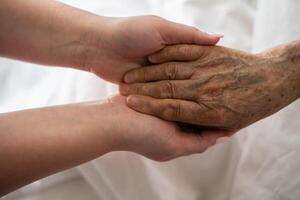 A young womans hands gently hold the hands of an elderly female patient in a hospital bed, symbolizing care, support, and compassion. photo