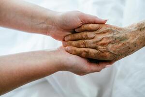 A young womans hands gently hold the hands of an elderly female patient in a hospital bed, symbolizing care, support, and compassion. photo