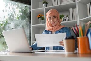 Smiling Muslim woman reviewing documents while working on a laptop in an office. photo