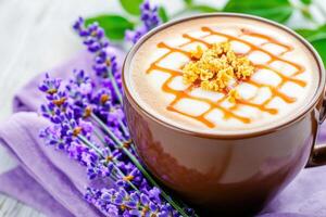 Close-up view of a latte topped with golden caramel drizzle, served in a mocha brown cup on a lavender napkin for a cozy aesthetic photo
