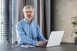 Confident professional man with glasses working on laptop in modern office setting. Workspace includes notebook, pen, tablet. Portrays productivity, focus, technology, and business environment. photo