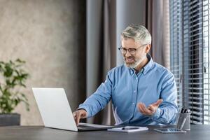 Senior businessman feeling frustrated due to malfunctioning laptop while sitting at modern office desk. Experiencing technical difficulties, visibly stressed. Surroundings include office plants, blinds, and stationary items. photo