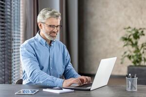 Mature man with grey hair and beard, working on a laptop in a home office environment, expressing confidence and productivity. photo