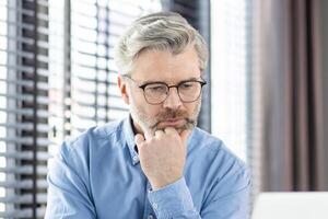 A mature businessman in glasses and a blue shirt is thinking deeply while working on a laptop in a modern office. The image conveys concentration, thoughtfulness, and dedication. photo