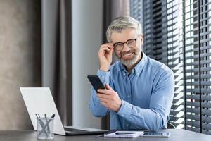 Smiling businessman sitting at desk using smartphone in modern office. Man working on laptop with notepad and glasses on table. Bright environment with window blinds in background photo